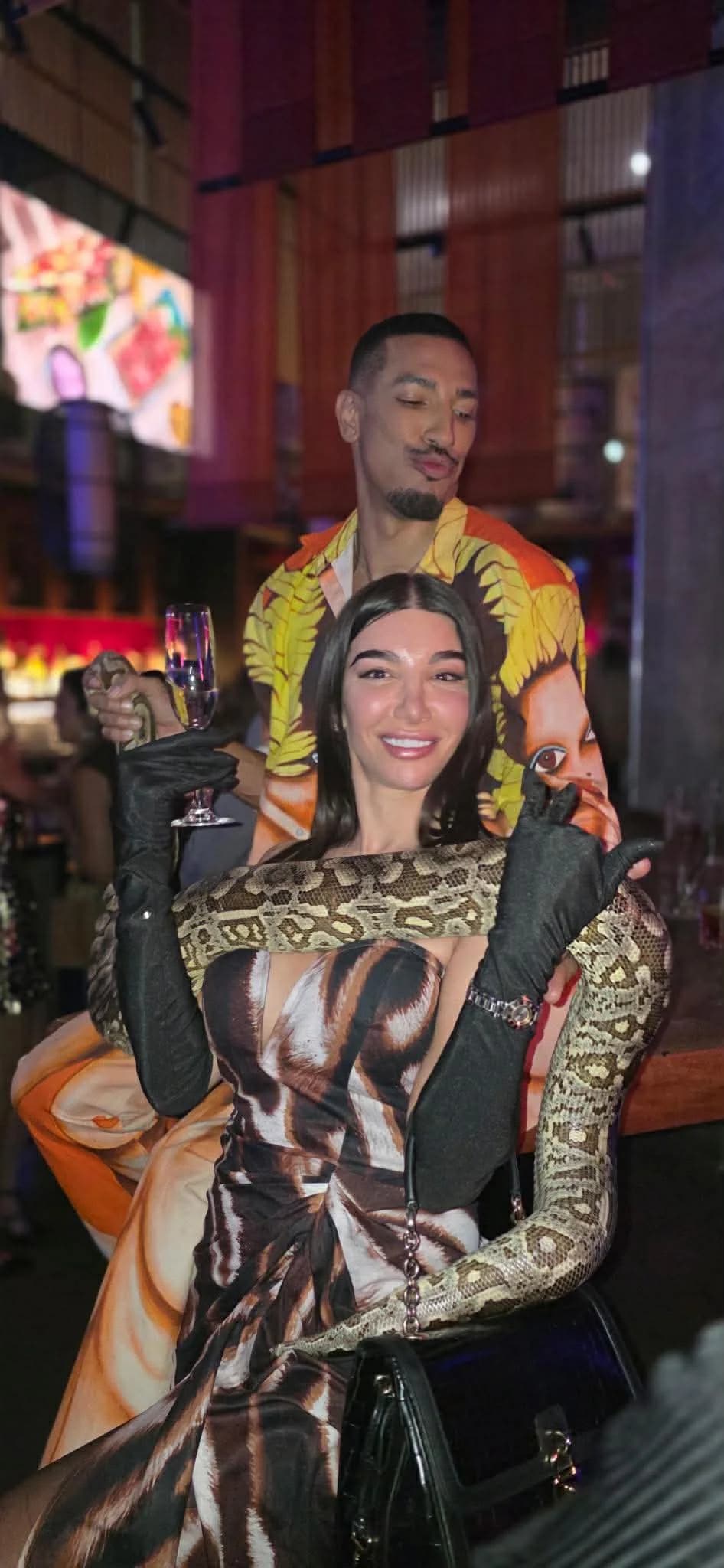 A guest in a brown patterned dress poses with a champagne flute and a python around her shoulders during a Toronto bar takeover