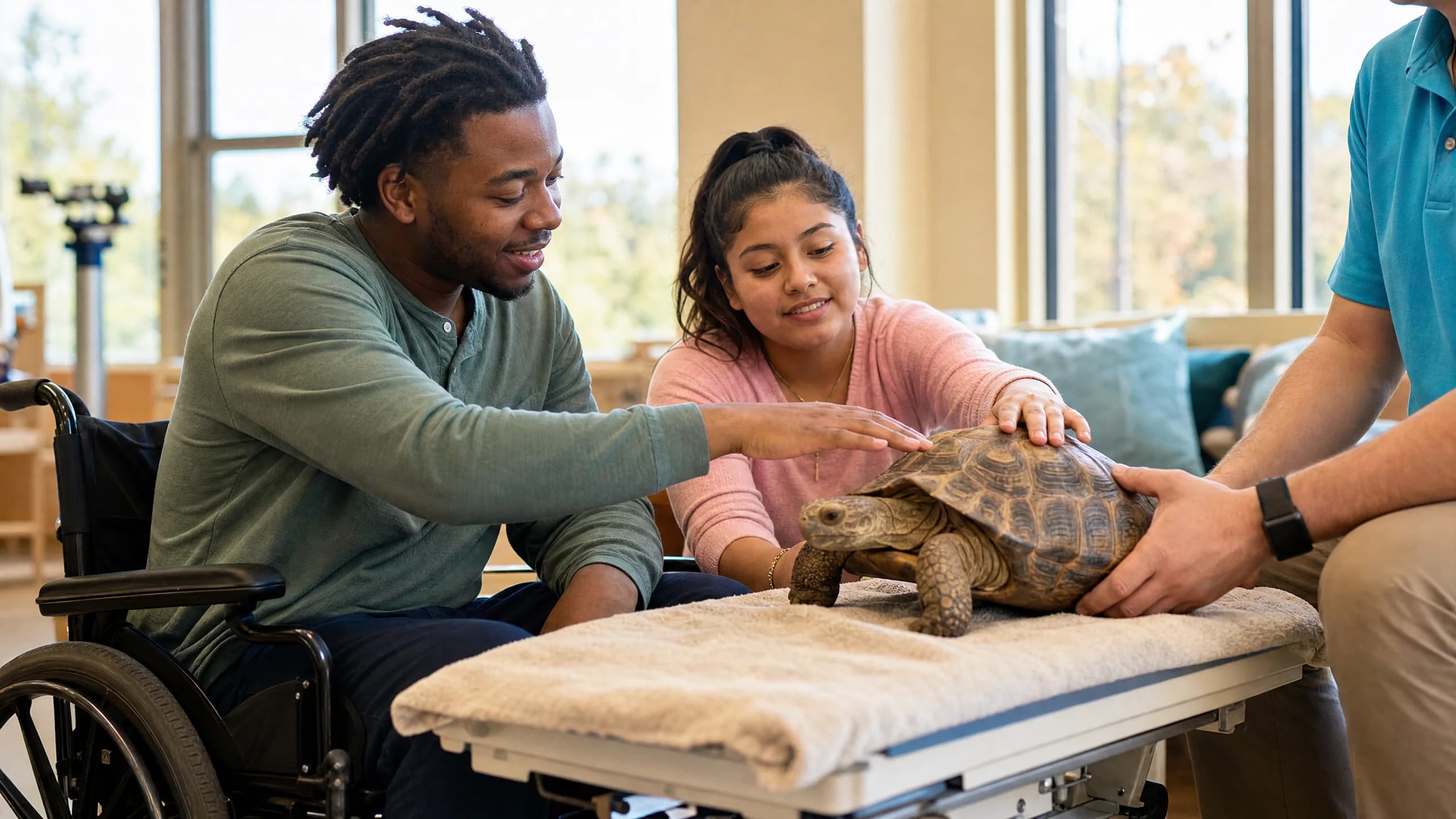A young Black adult using a wheelchair and a Latina teen seated alongside both gently touching a tortoise resting on a height-adjusted padded table in a bright accessible community room