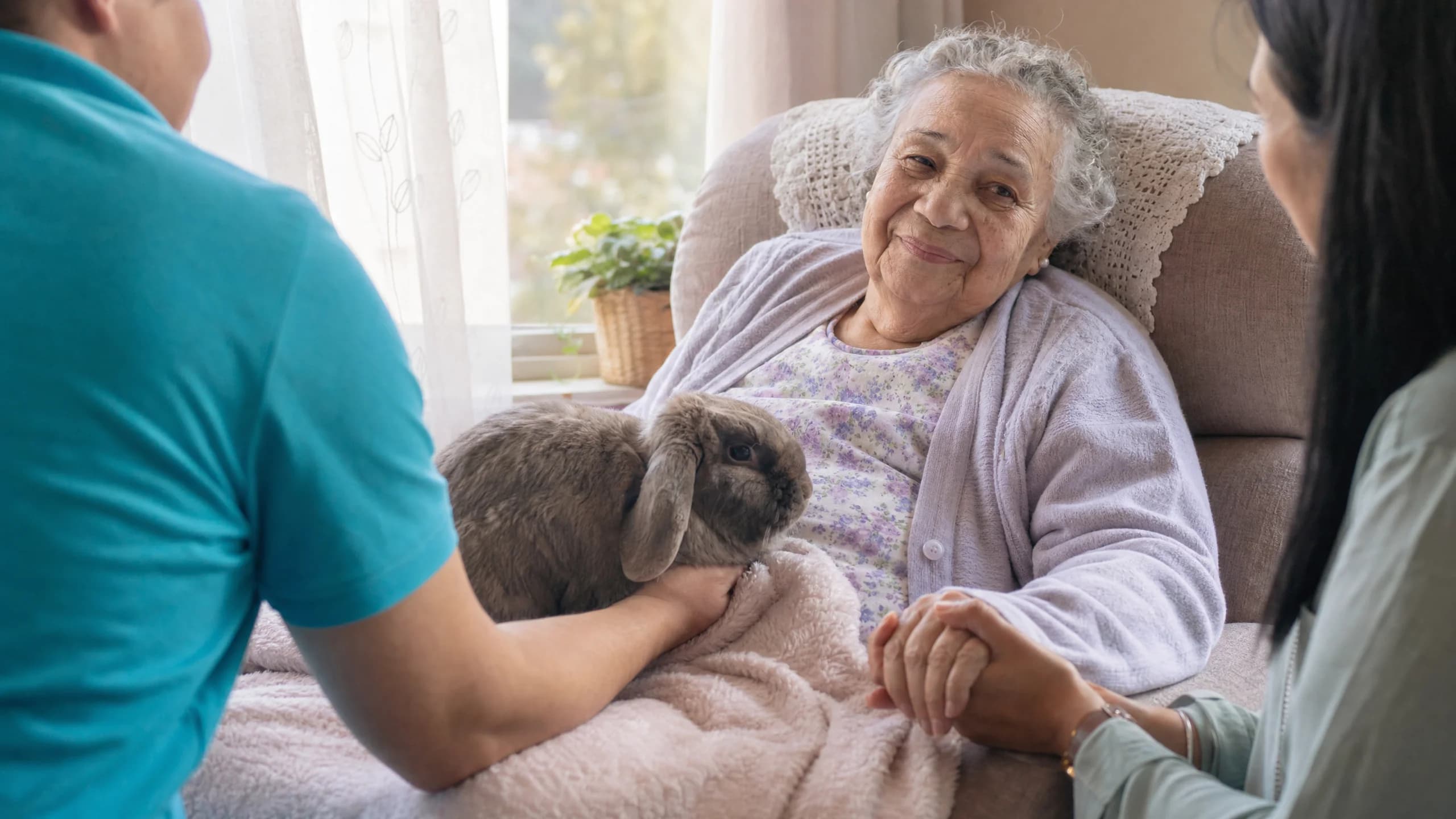 An elderly woman resting comfortably in a sunlit care-home armchair smiles softly as a calm lop-eared rabbit rests on a soft blanket on her lap, with a family member gently holding her hand and a Hands On Exotics handler in a cyan polo at the edge of the frame