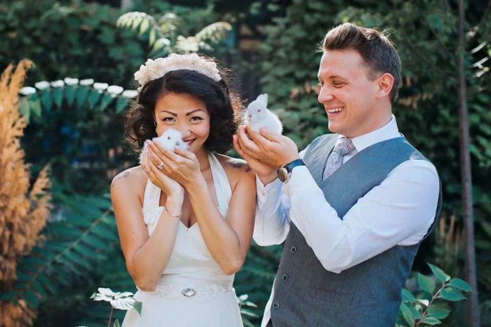 A bride in a white halter dress and floral crown laughs with her groom in a grey vest as they each cradle a tiny white baby rabbit at a sunlit garden wedding