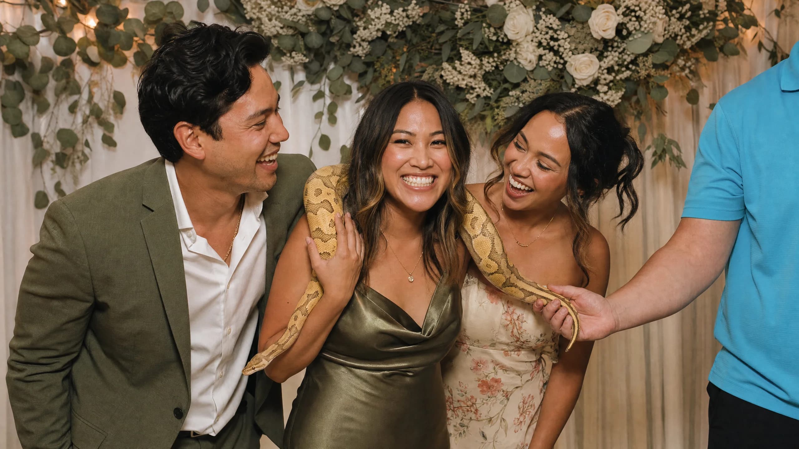 Three wedding guests in cocktail attire posed shoulder-to-shoulder at a photo booth, one woman in a green dress laughing with a calm ball python draped across her shoulders, in front of a eucalyptus and white floral backdrop